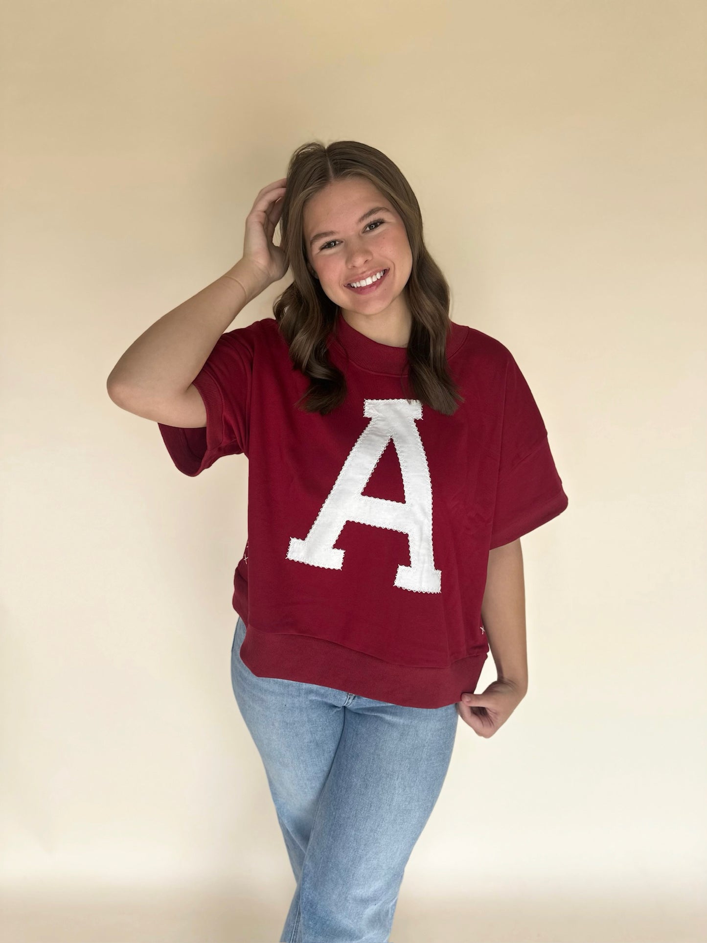 Woman wearing a crimson oversized short-sleeve top with a large white varsity-style A on the front, paired with light wash jeans, standing against a neutral backdrop.