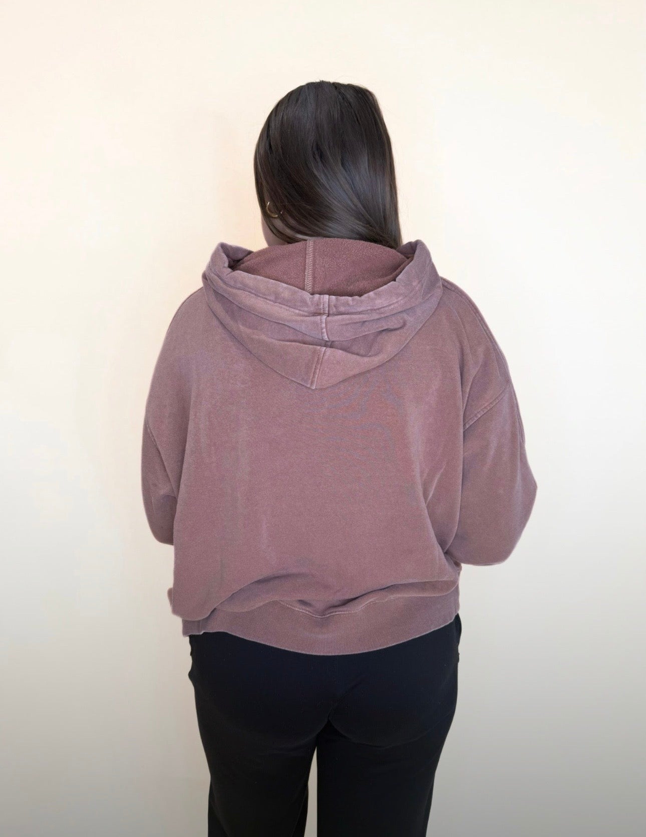 Back view of a woman wearing a faded mocha brown hoodie with a relaxed fit and black pants, hair down, shown against a light neutral background.