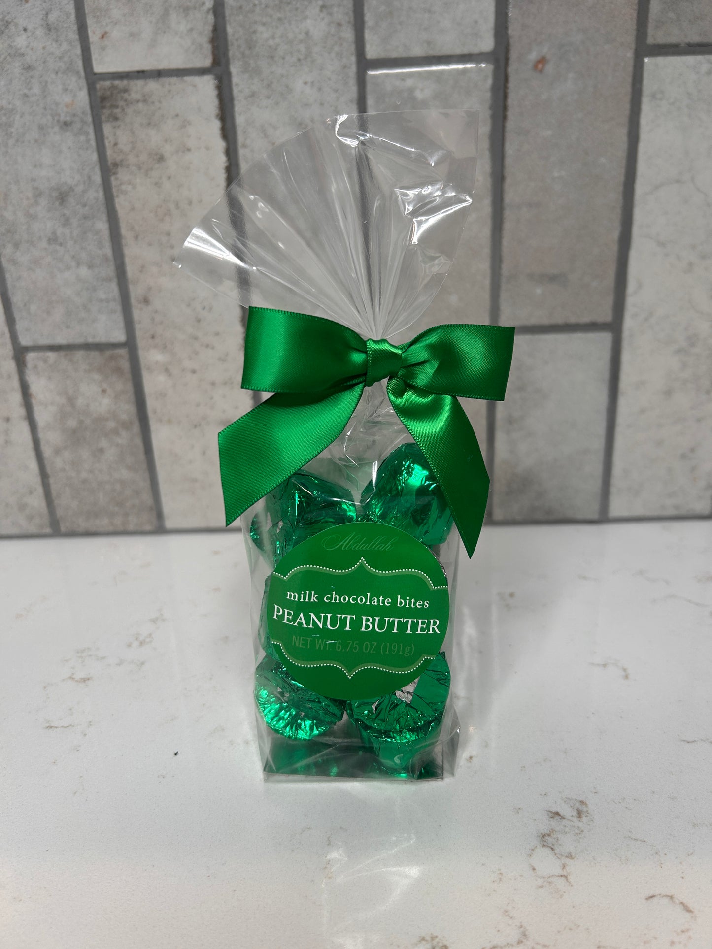 A clear gift bag filled with green-foil wrapped milk chocolate peanut butter bites, tied with a green satin bow, displayed on a white countertop with a gray tile background.