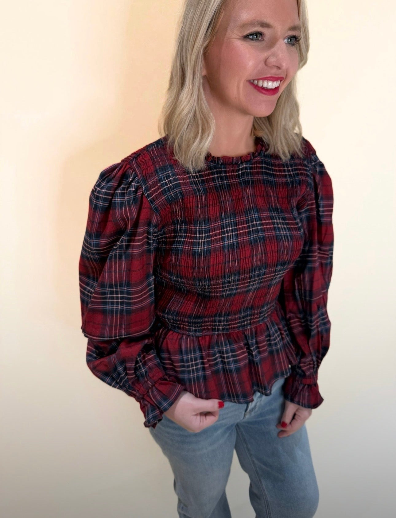 Close angle of woman wearing a red and navy plaid peplum blouse with gathered sleeves and smocked fit, styled casually with denim jeans.