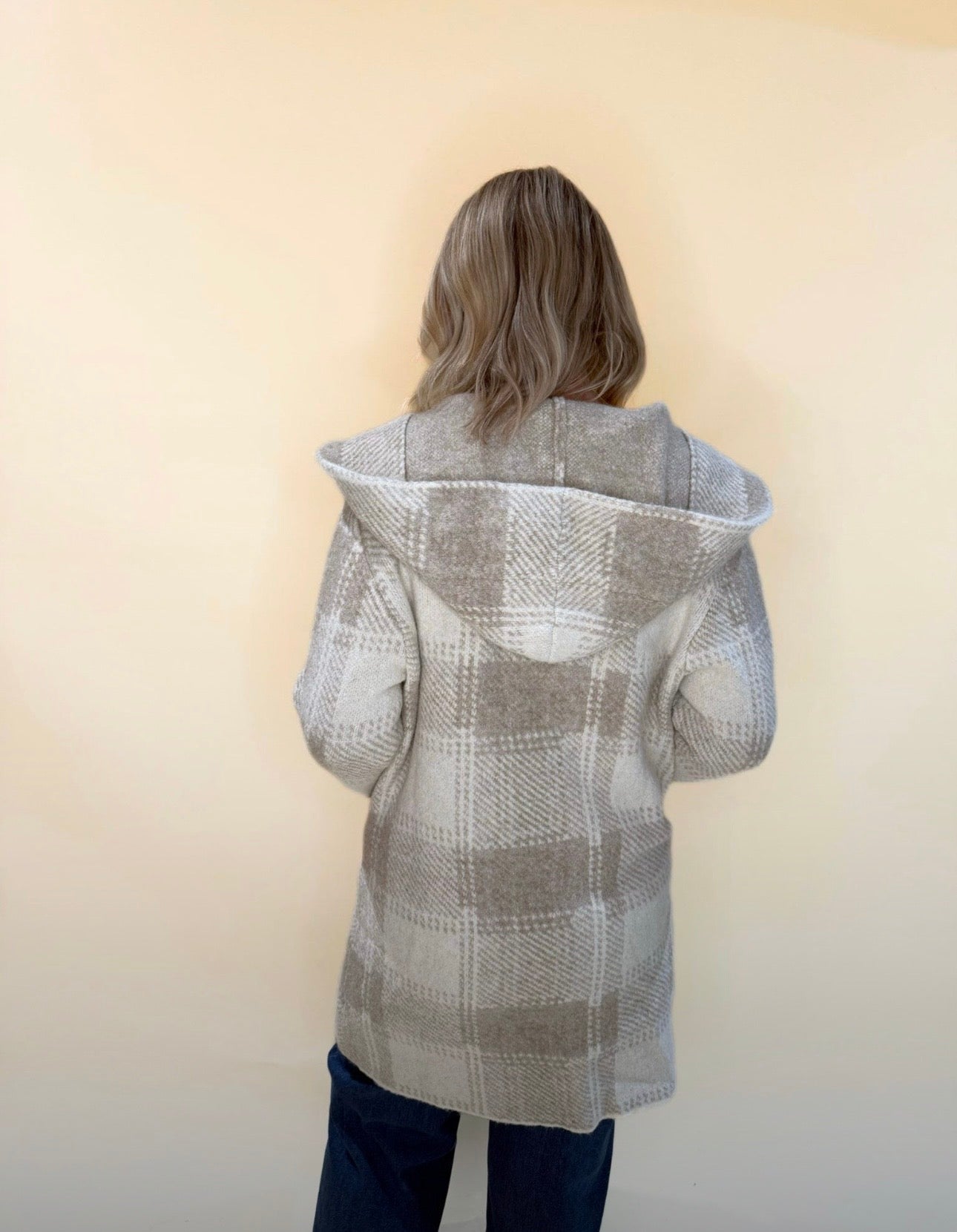 Back view of a taupe and cream plaid knit hooded cardigan, showing relaxed fit and draped hood, styled with blue denim.