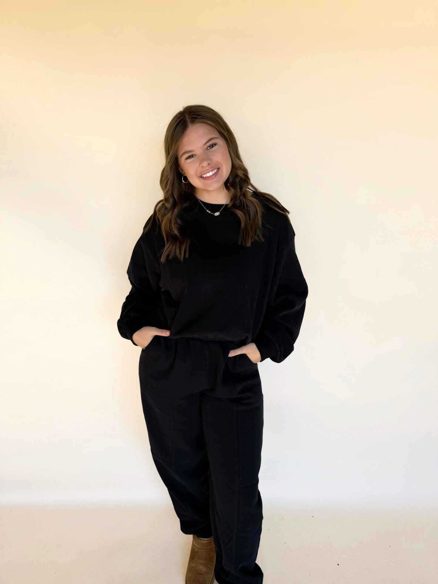 Woman wearing black matching lounge set with long sleeve top and jogger pants, hands in pockets, smiling against neutral backdrop.