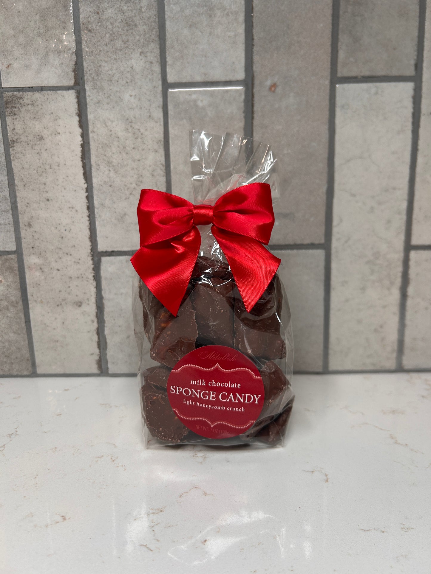 A clear gift bag filled with milk chocolate sponge candy pieces, tied with a red satin bow, displayed on a white countertop with a gray tile background.