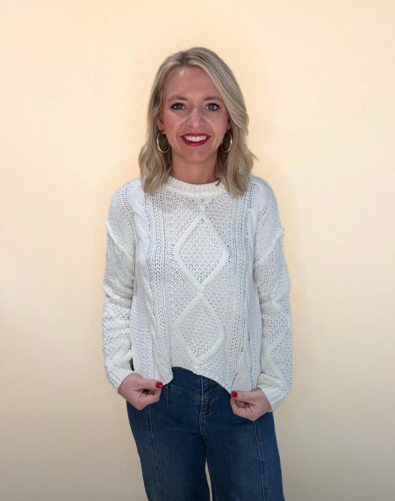 Front angle view of white cable knit sweater with diamond knit pattern and blue jeans, model smiling against a soft cream backdrop.