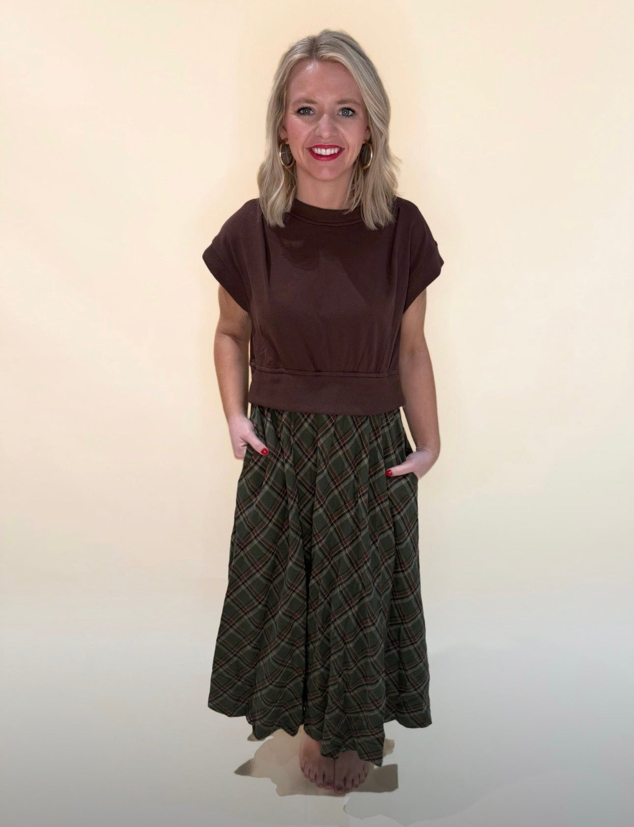 Full body view of brown short-sleeve top styled with a flowy green plaid maxi skirt, hands in pockets, smiling at camera against a cream backdrop.