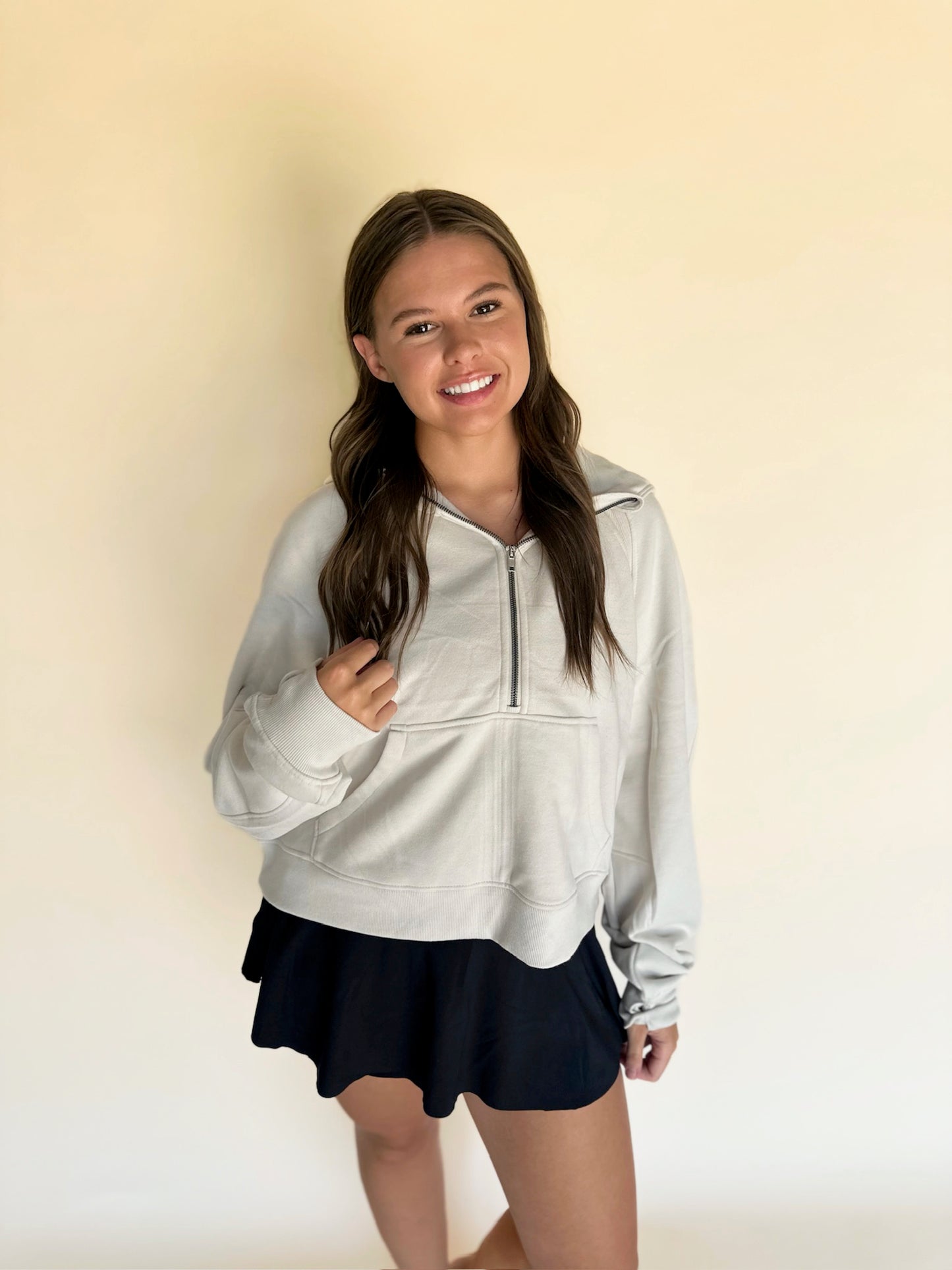 Woman wearing a light gray zip-up sweatshirt and black skirt against a beige background
