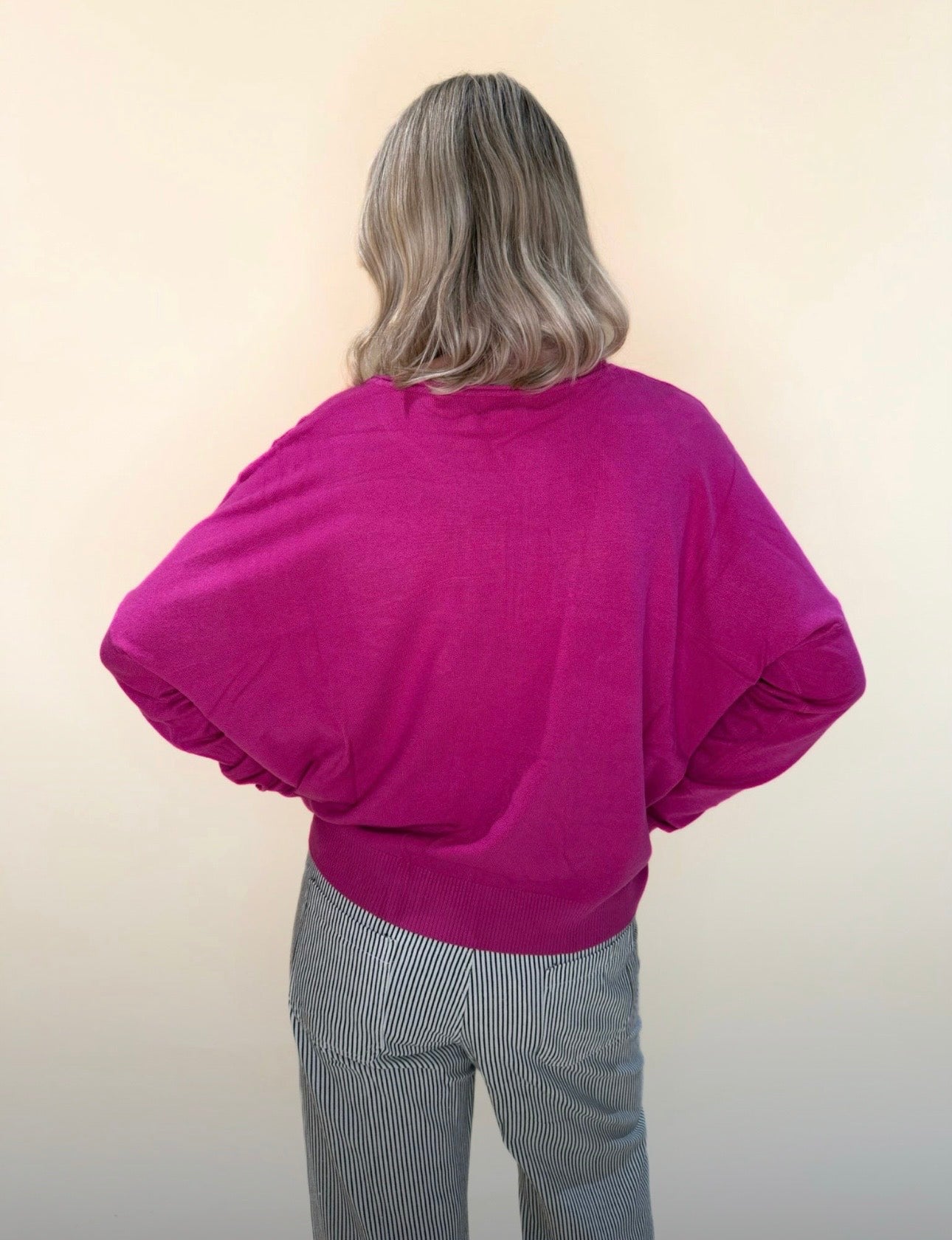 Back view of a woman wearing a fuchsia sweater with relaxed sleeves and black and white striped pants, standing against a soft cream background.