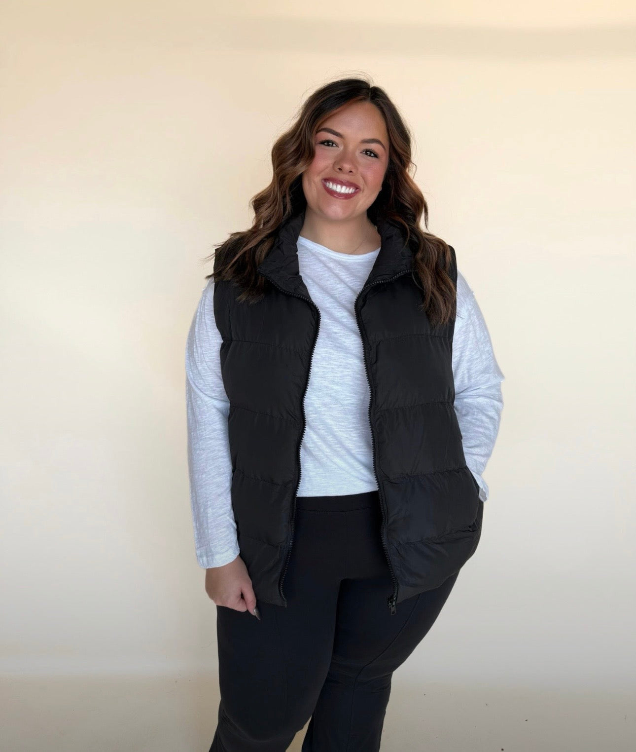 Woman wearing a black puffer vest over a white long-sleeve top and black leggings, standing and smiling in front of a minimal neutral background.