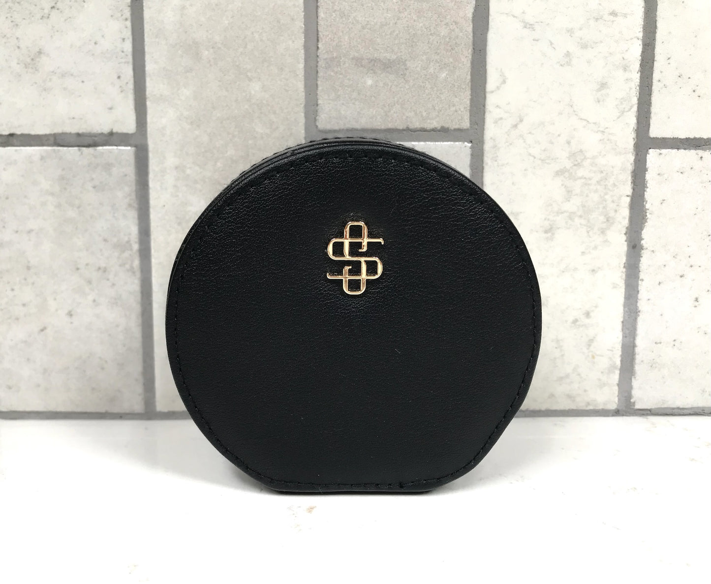 Black round jewelry case with a gold logo on a light gray tiled background
