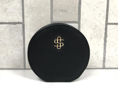 Black round jewelry case with a gold logo on a light gray tiled background