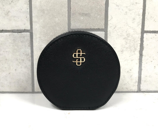 Black round jewelry case with a gold logo on a light gray tiled background