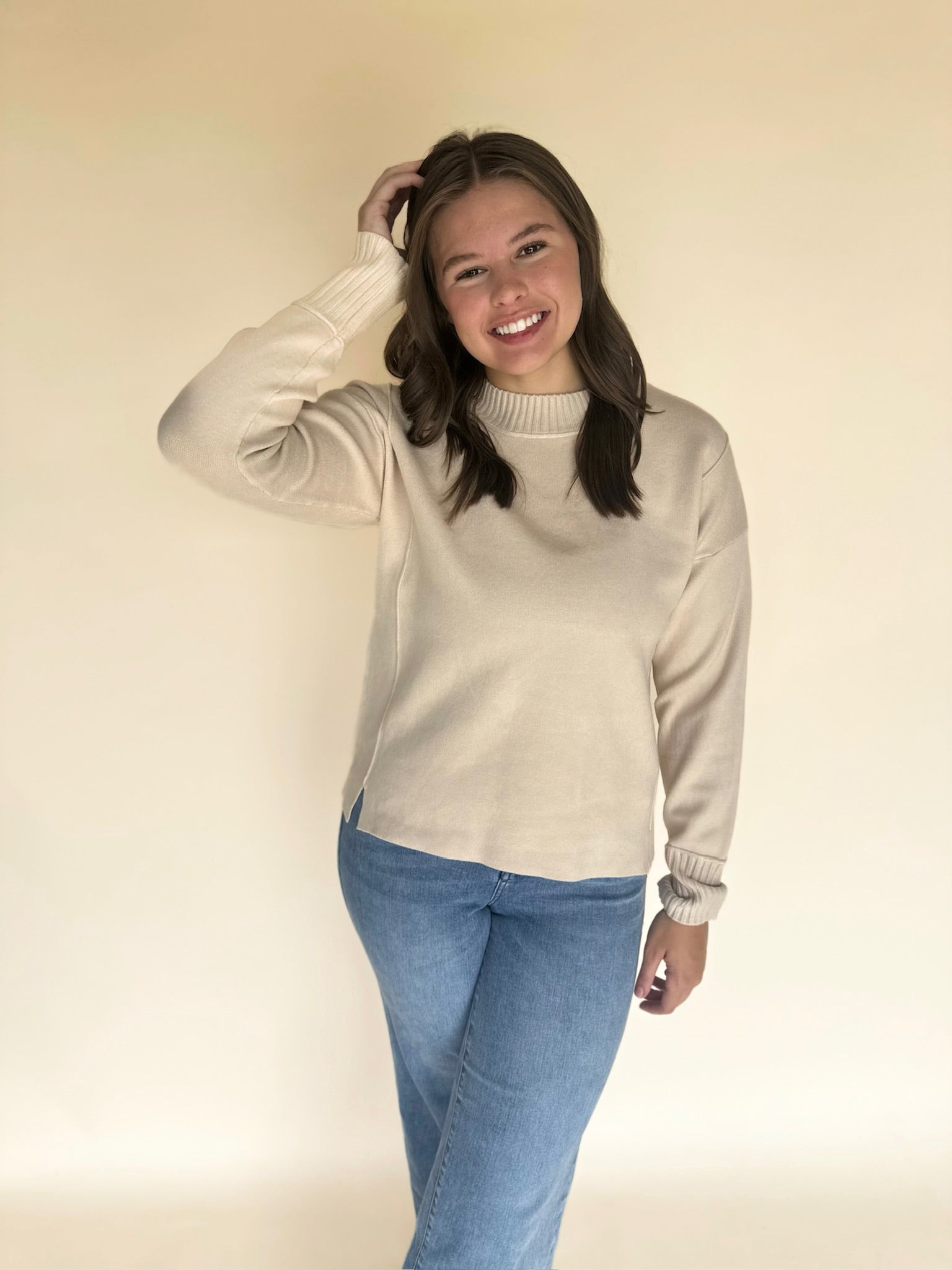 Woman wearing an oatmeal sweater and blue jeans against a plain background