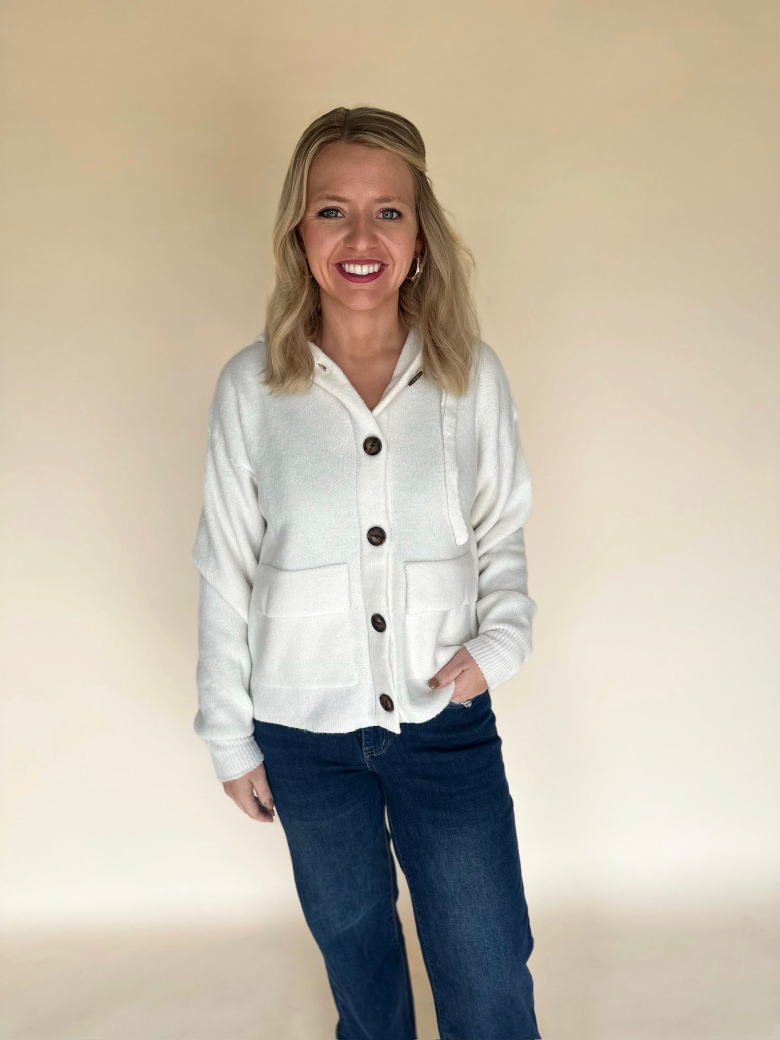Woman in a cream knit button-front hoodie cardigan and dark denim jeans standing against a neutral background, showcasing a casual cozy outfit.
