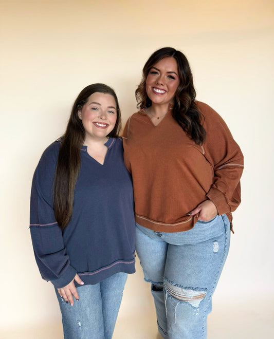 Two women wearing casual long-sleeve tops; one in a navy V-neck pullover with exposed contrast stitching and the other in a rust V-neck top with seam detailing, both styled with distressed jeans against a neutral background.