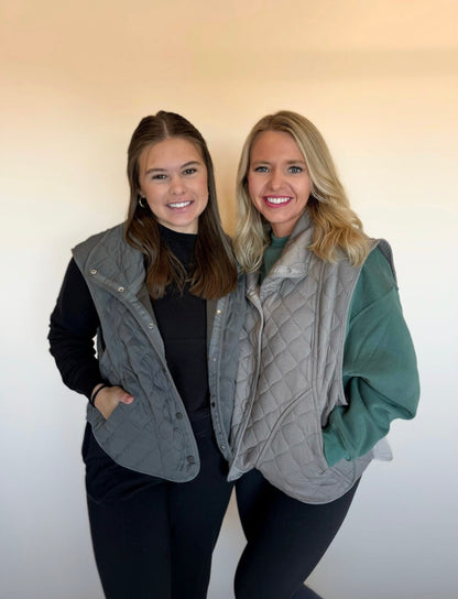 Two women wearing gray quilted vests against a plain background