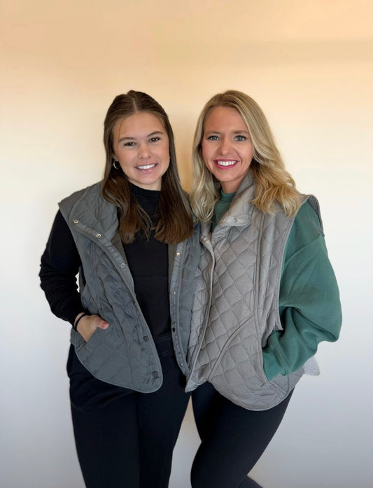 Two women wearing gray quilted vests against a plain background