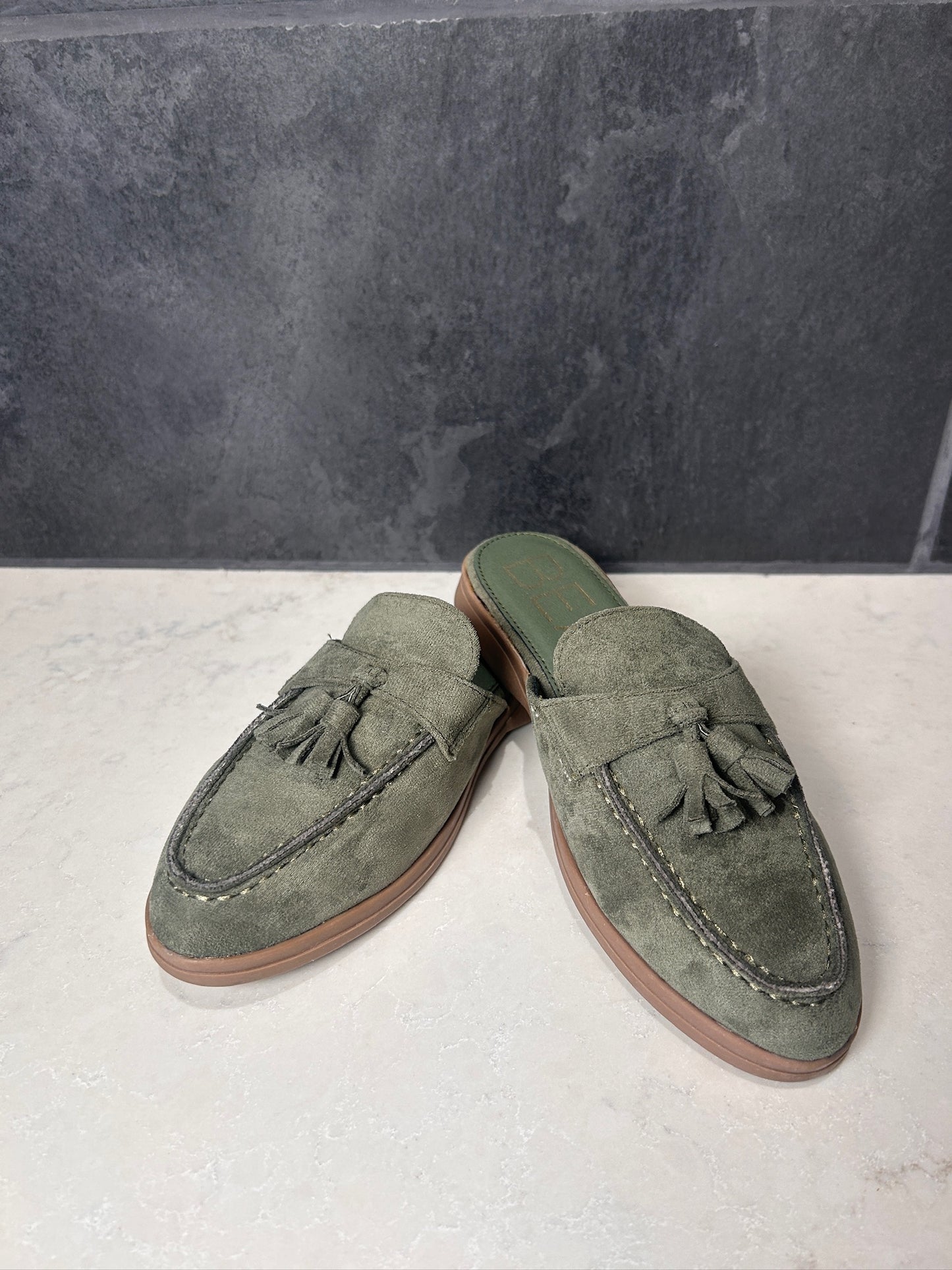 Pair of green suede loafers with tassels on a light gray surface.