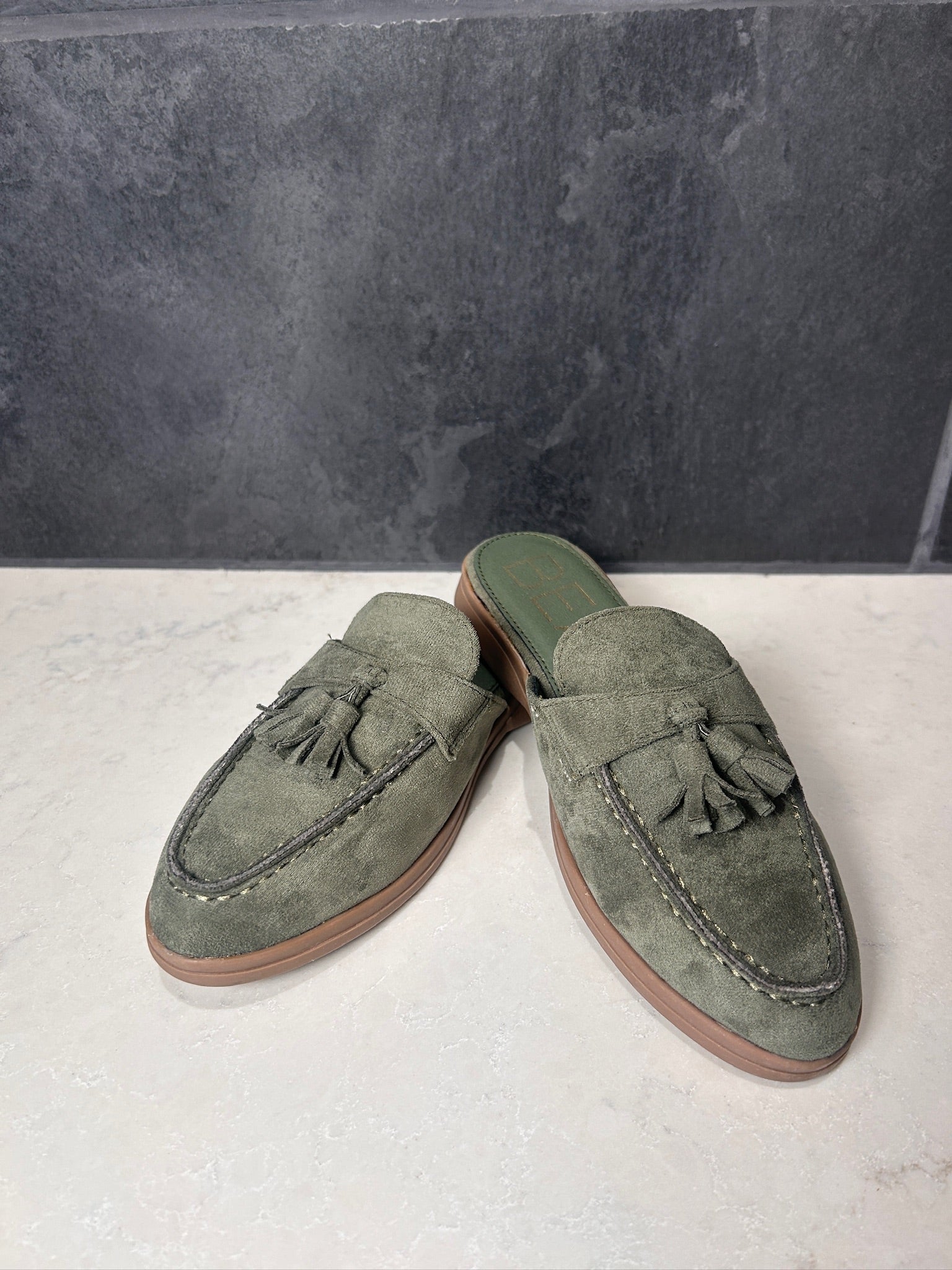 Pair of green suede loafers with tassels on a light gray surface.