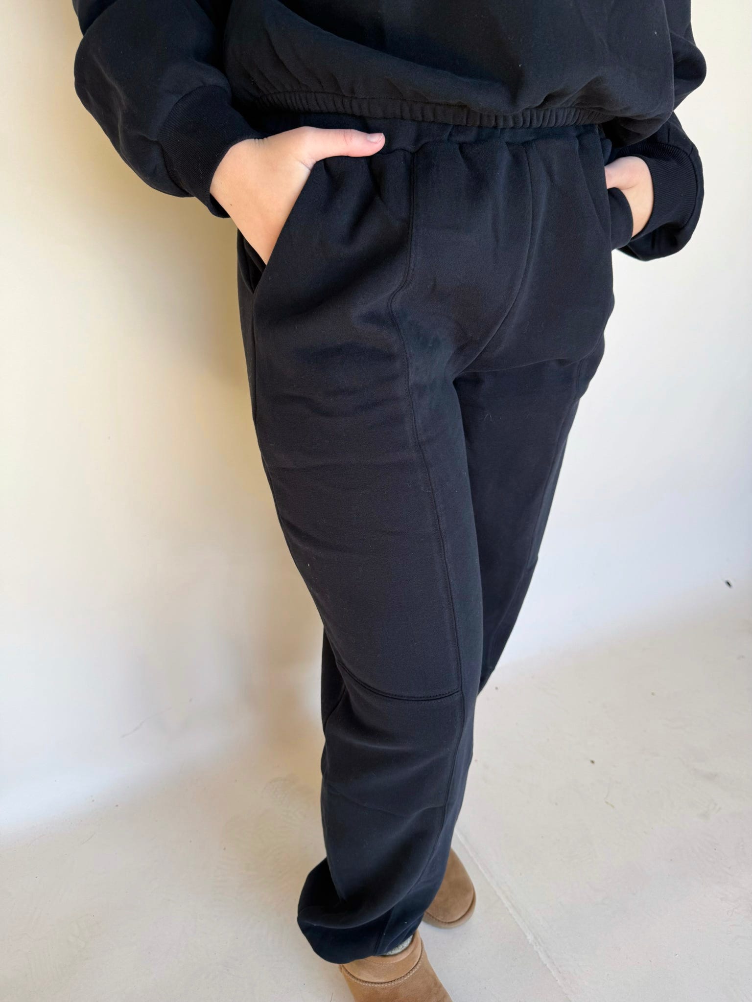 Black high-waisted jogger pants with elastic waistband and pockets, styled with hands tucked in, relaxed cozy fit.