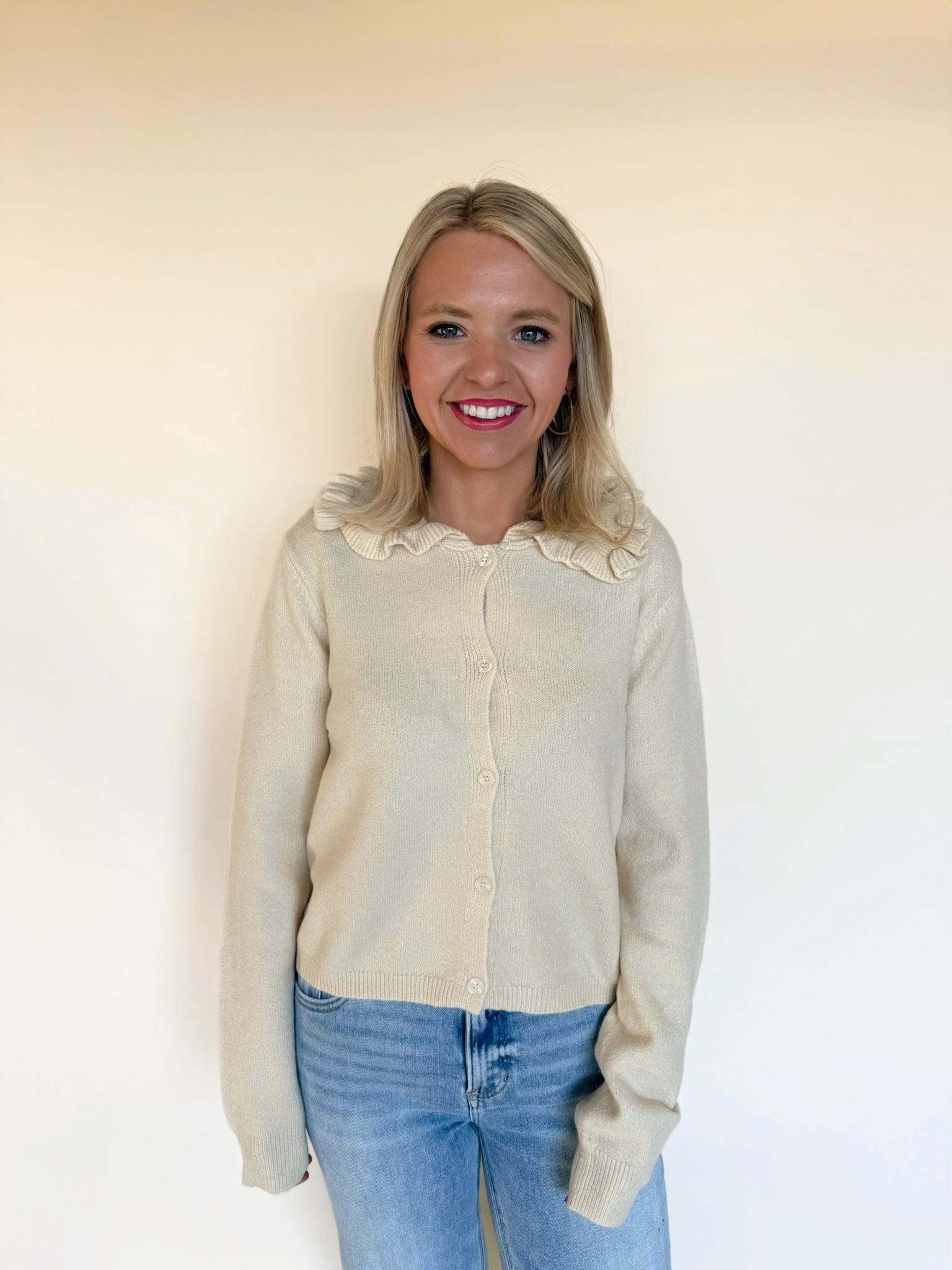 Woman wearing a cream button-front cardigan with a ruffled collar paired with light-wash blue jeans, standing against a neutral backdrop.