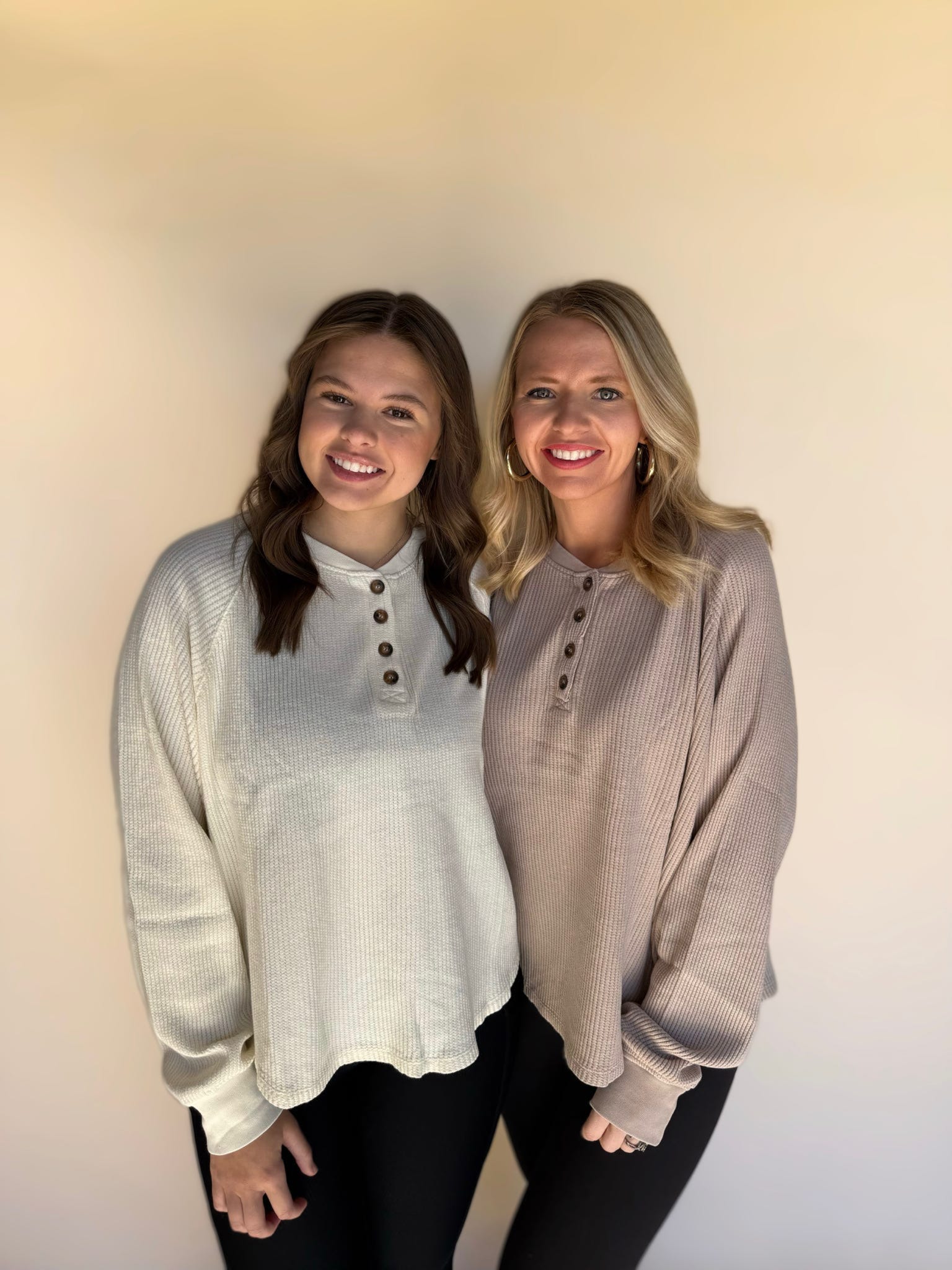 Two women wearing light-colored long-sleeve shirts standing against a plain background