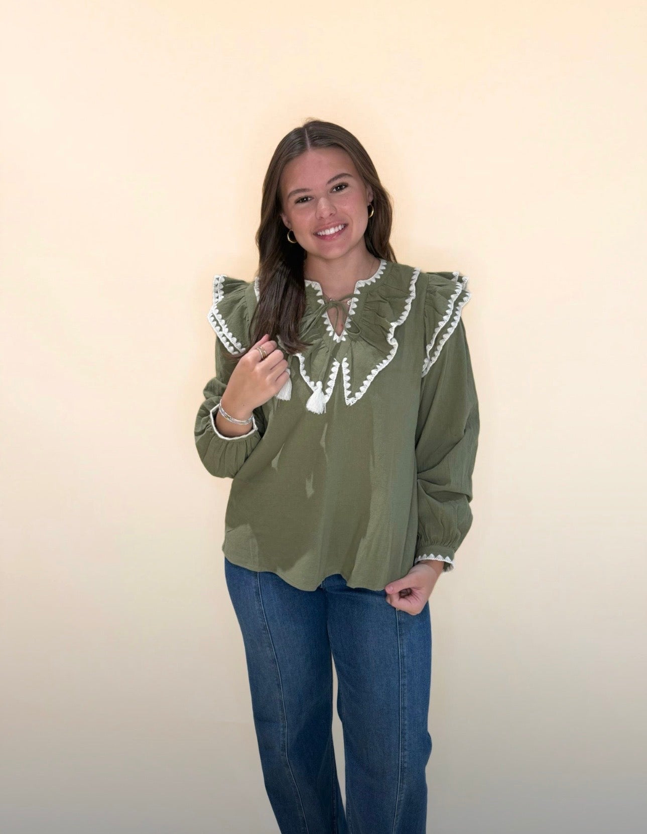 Long sleeve olive green ruffle blouse with cream embroidered detail and tassel neck tie, worn with dark wash jeans.