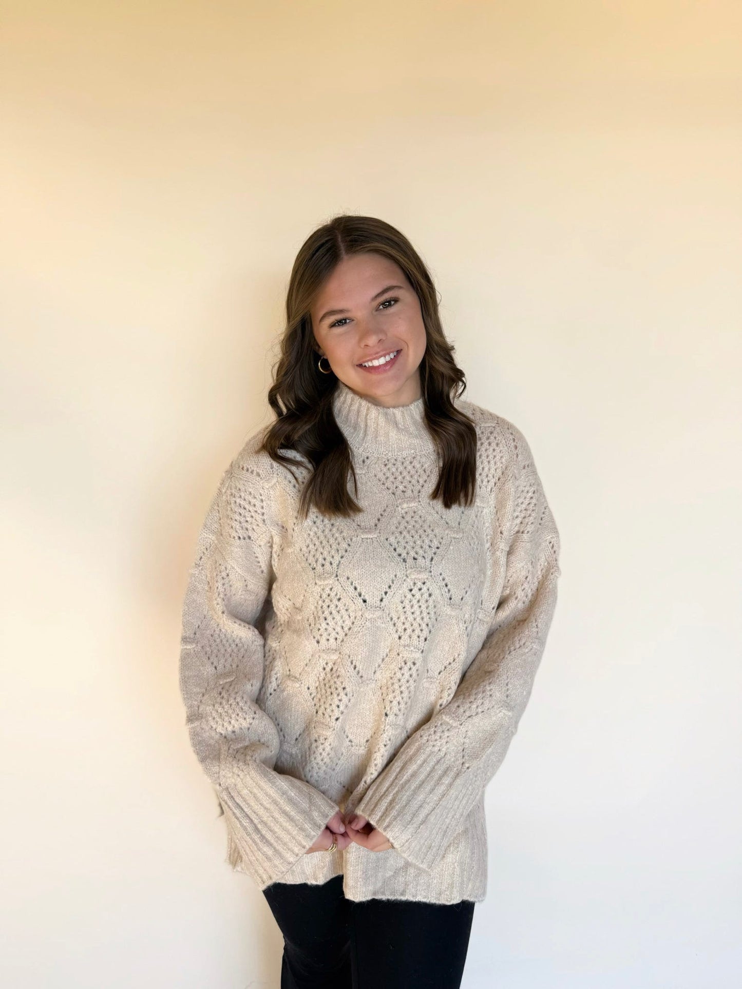 Woman wearing a cream textured knit sweater featuring a diamond pattern and a mock neck, styled with black leggings against a soft neutral backdrop.