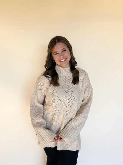 Woman wearing a cream textured knit sweater featuring a diamond pattern and a mock neck, styled with black leggings against a soft neutral backdrop.