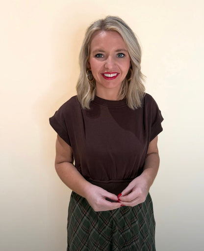 Close-up front view of a woman in a brown short-sleeve top and green plaid skirt, smiling with hands together, styled against a soft cream backdrop.