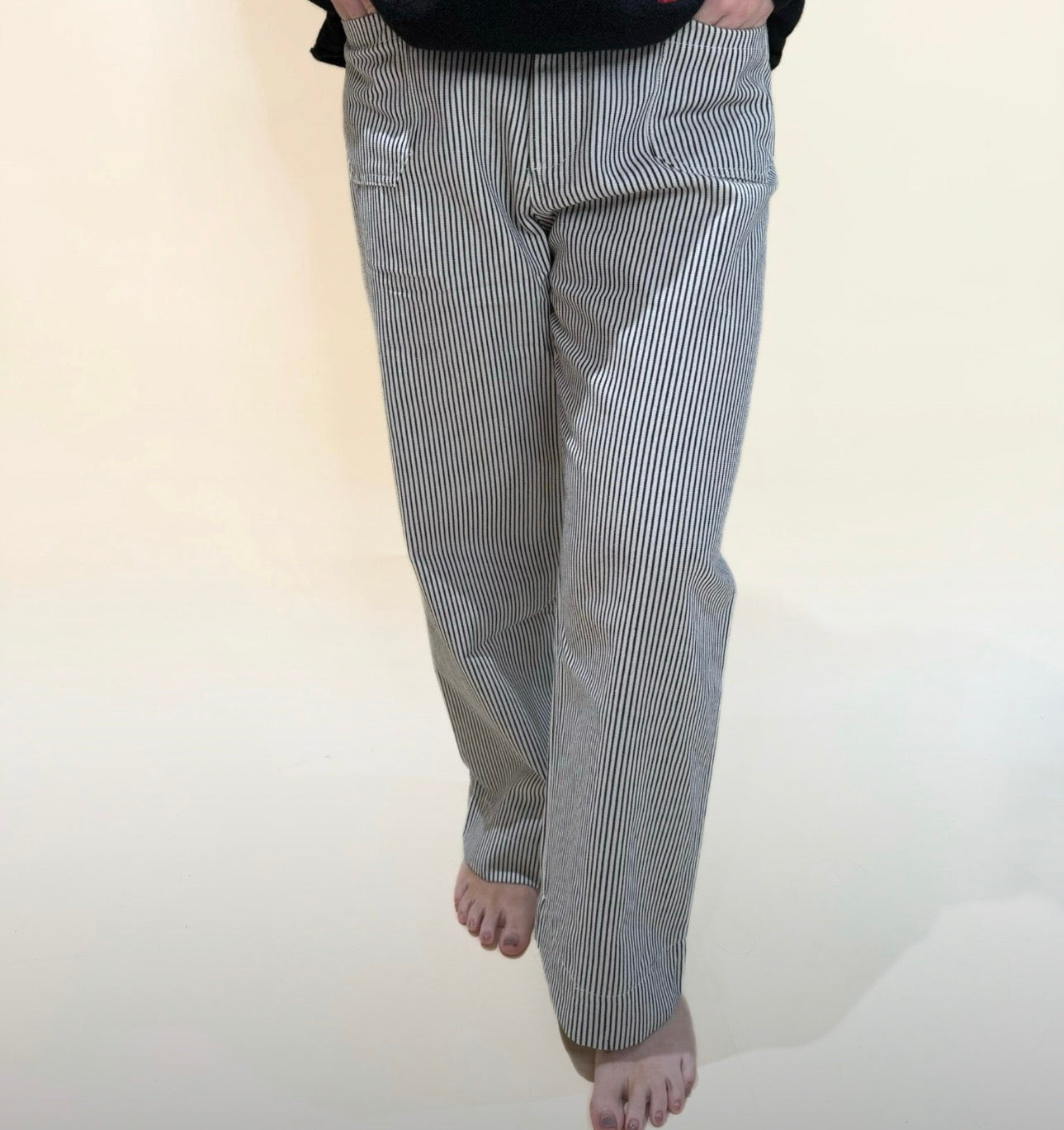 Front view of a person wearing black and white striped straight-leg pants, hands in pockets, barefoot, standing against a neutral light background.