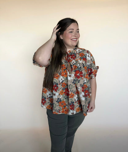 Woman wearing a taupe floral babydoll top with rust, navy, and white flowers, paired with dark pants. Short puff sleeves and tie-neck detail. Square 1 Boutique styling.