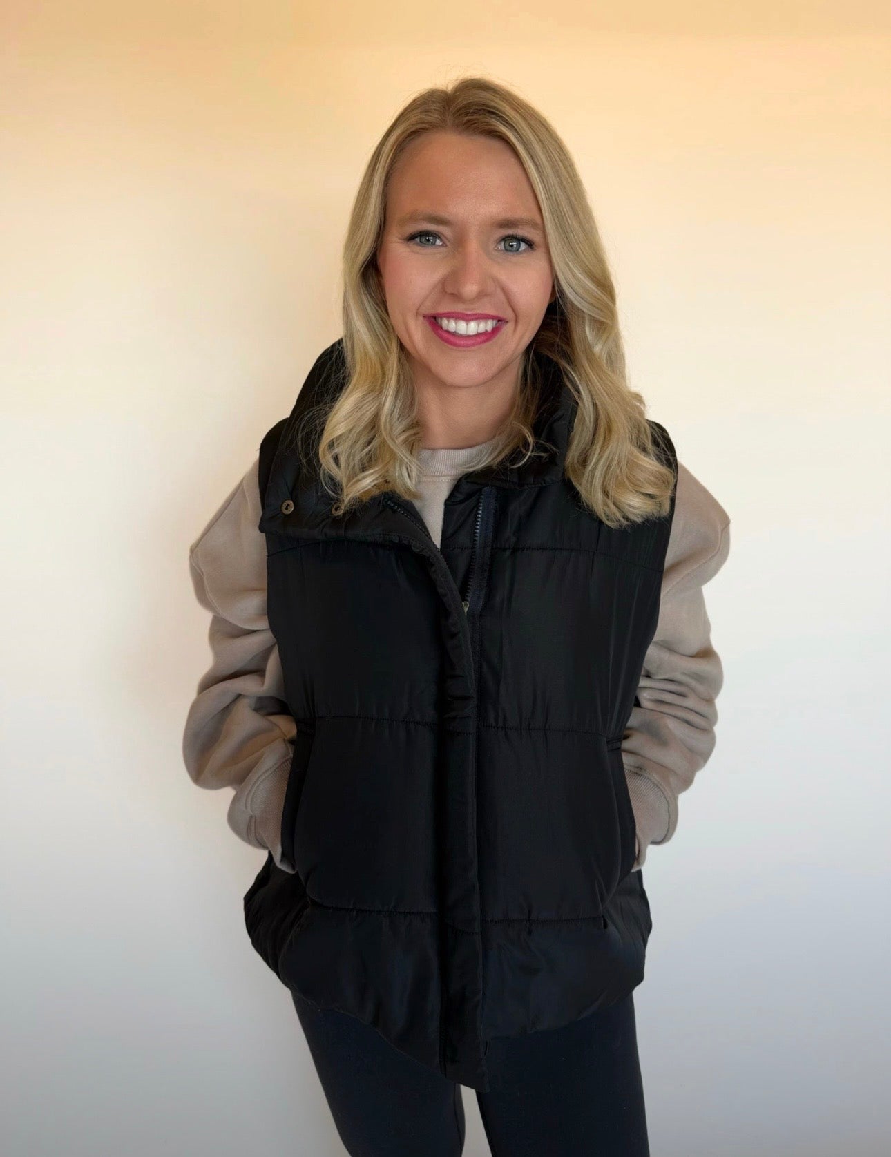 Woman wearing a black puffer vest over a beige jacket against a plain background