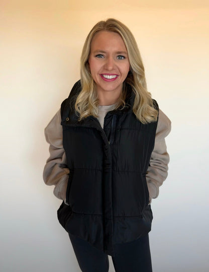 Woman wearing a black puffer vest over a beige jacket against a plain background