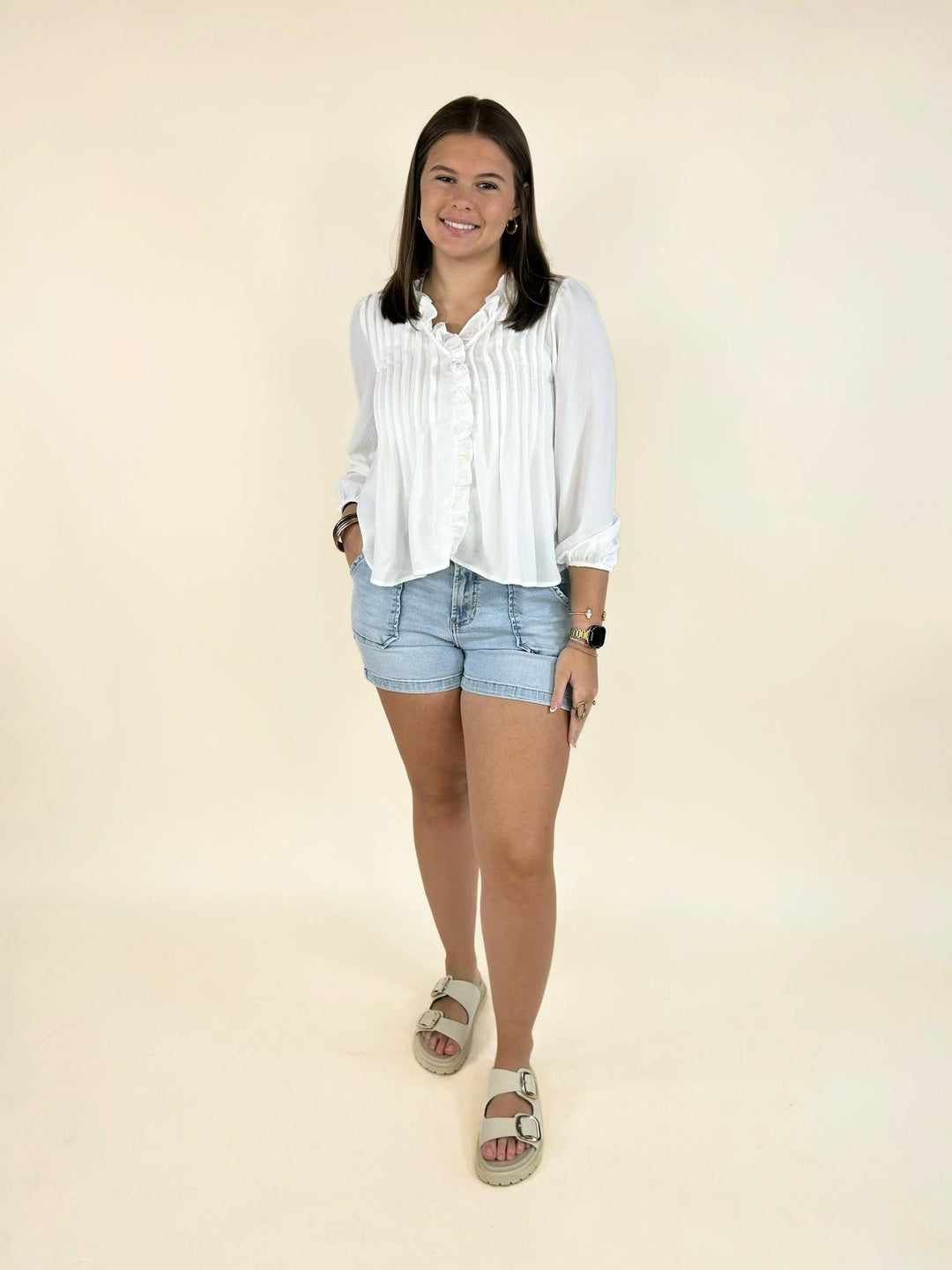 Front view of a white ruffle trim pintuck blouse styled with light wash denim shorts and sandals.