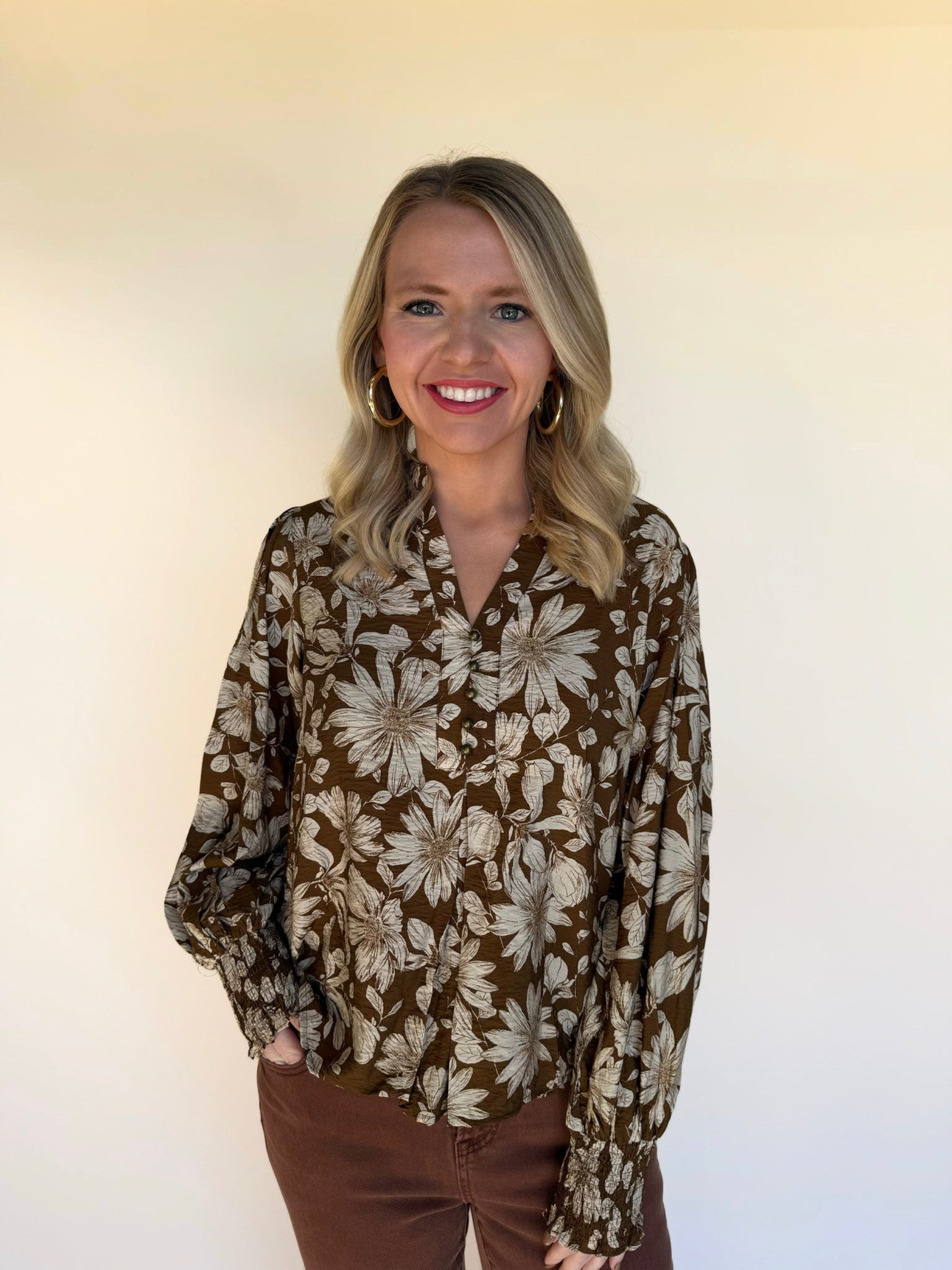 Close-up front view of a woman in a brown floral print blouse with long smocked sleeves and gold hoop earrings.