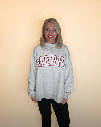 Woman wearing oversized light gray sweatshirt with red “MERRY” lettering, styled with black leggings on neutral backdrop.