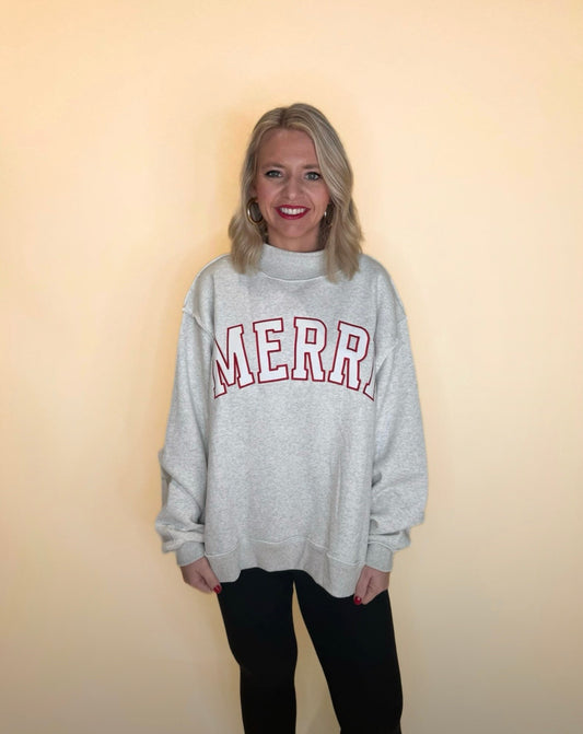Woman wearing oversized light gray sweatshirt with red “MERRY” lettering, styled with black leggings on neutral backdrop.