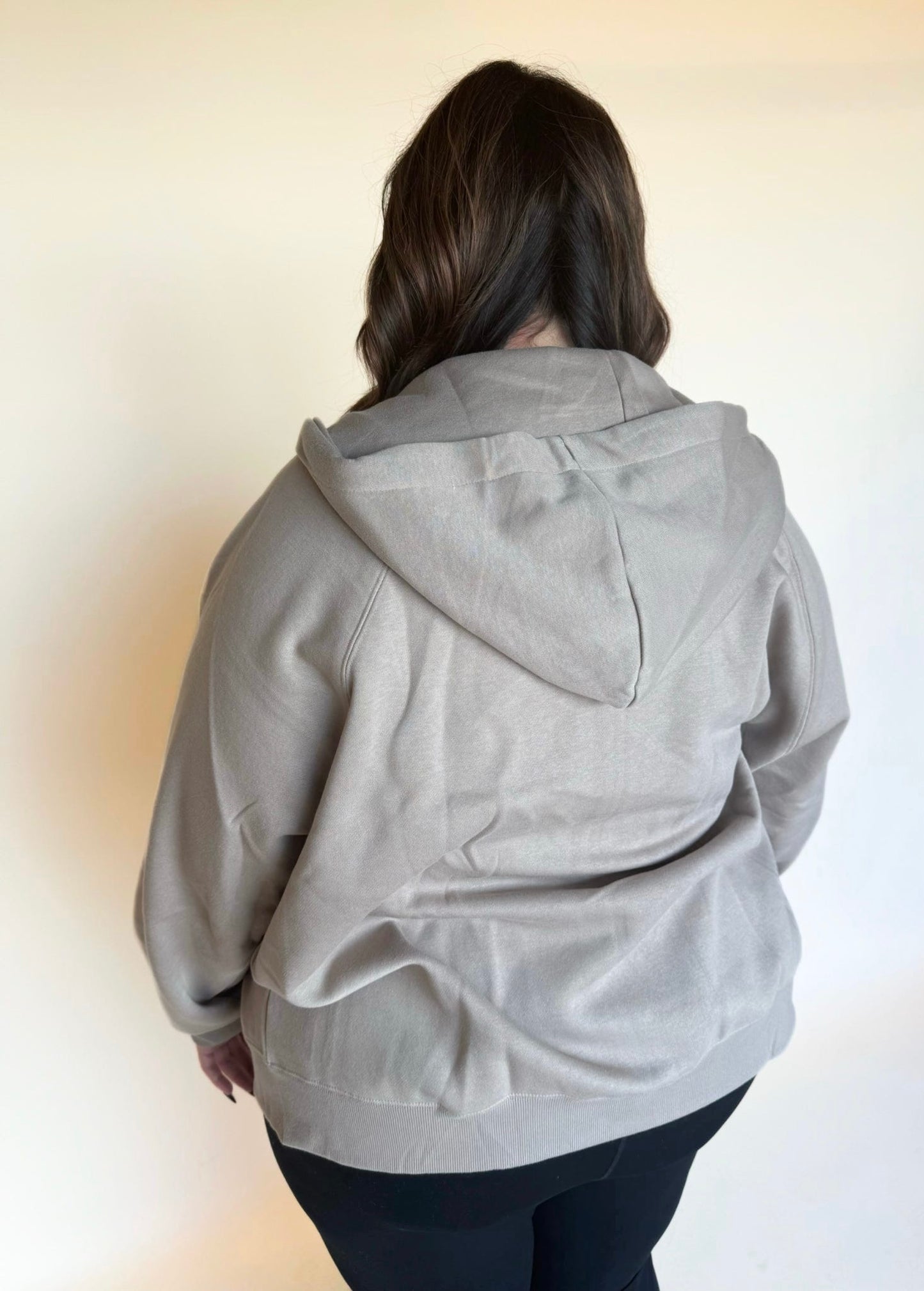 Back view of a woman wearing a taupe zip-up hoodie, highlighting the hood and casual relaxed fit.