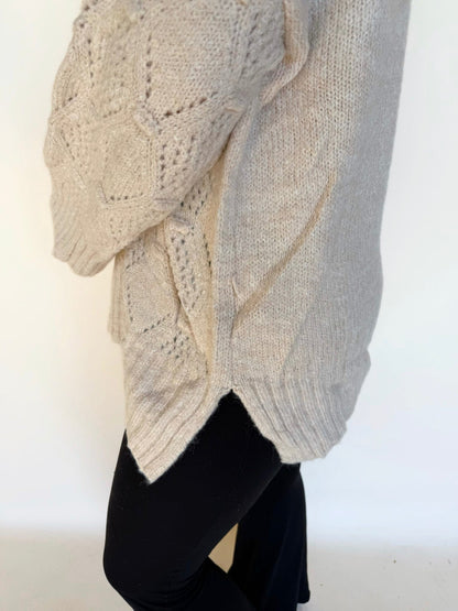 Close-up side view of a cream textured knit sweater highlighting the diamond pattern and side slit hem, styled with black leggings.