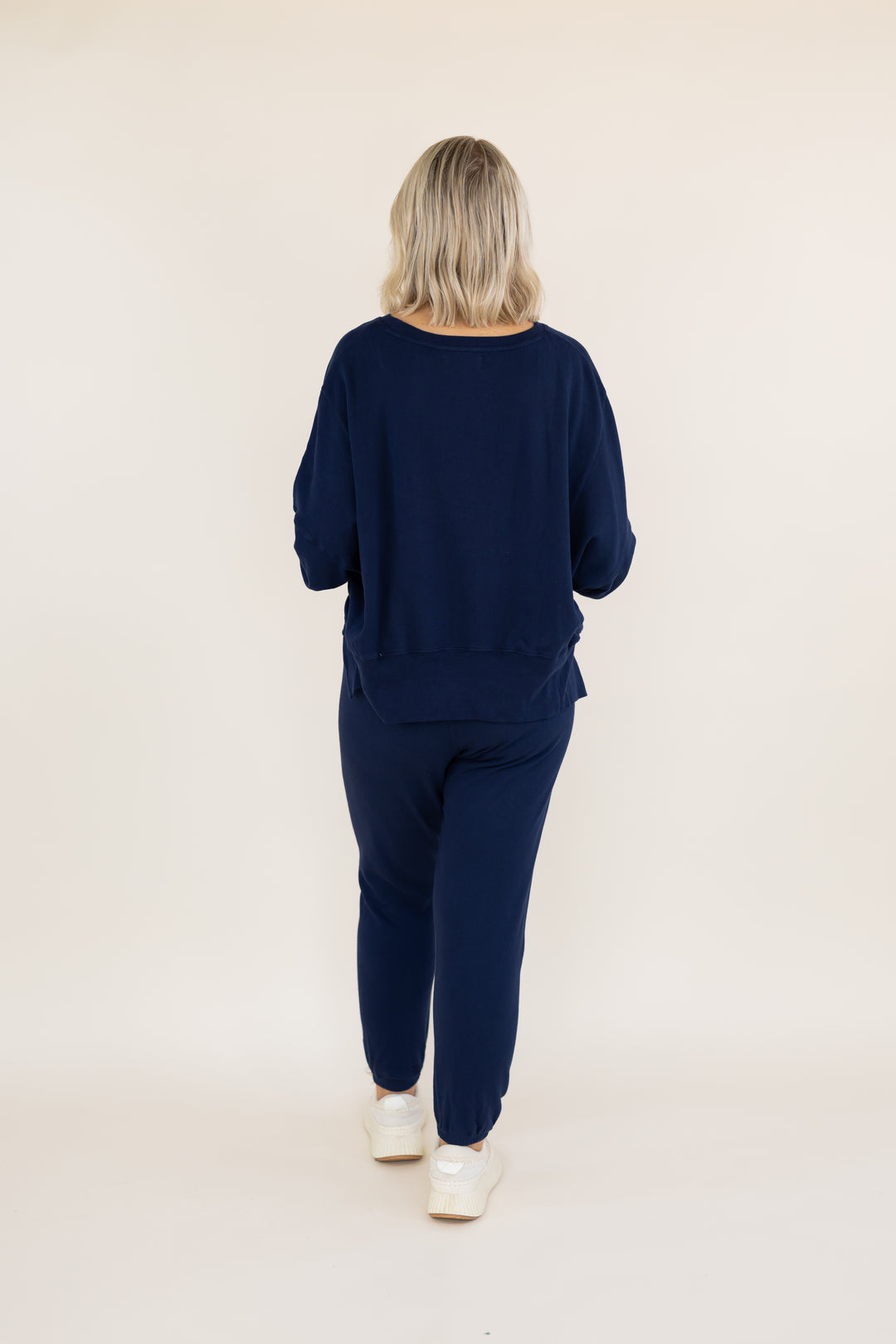 A woman viewed from behind wearing a matching navy blue lounge set consisting of a long-sleeve top and jogger pants, paired with white sneakers, against a plain off-white studio background.