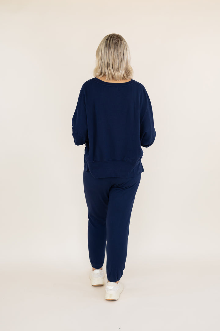 A woman viewed from behind wearing a matching navy blue lounge set consisting of a long-sleeve top and jogger pants, paired with white sneakers, against a plain off-white studio background.