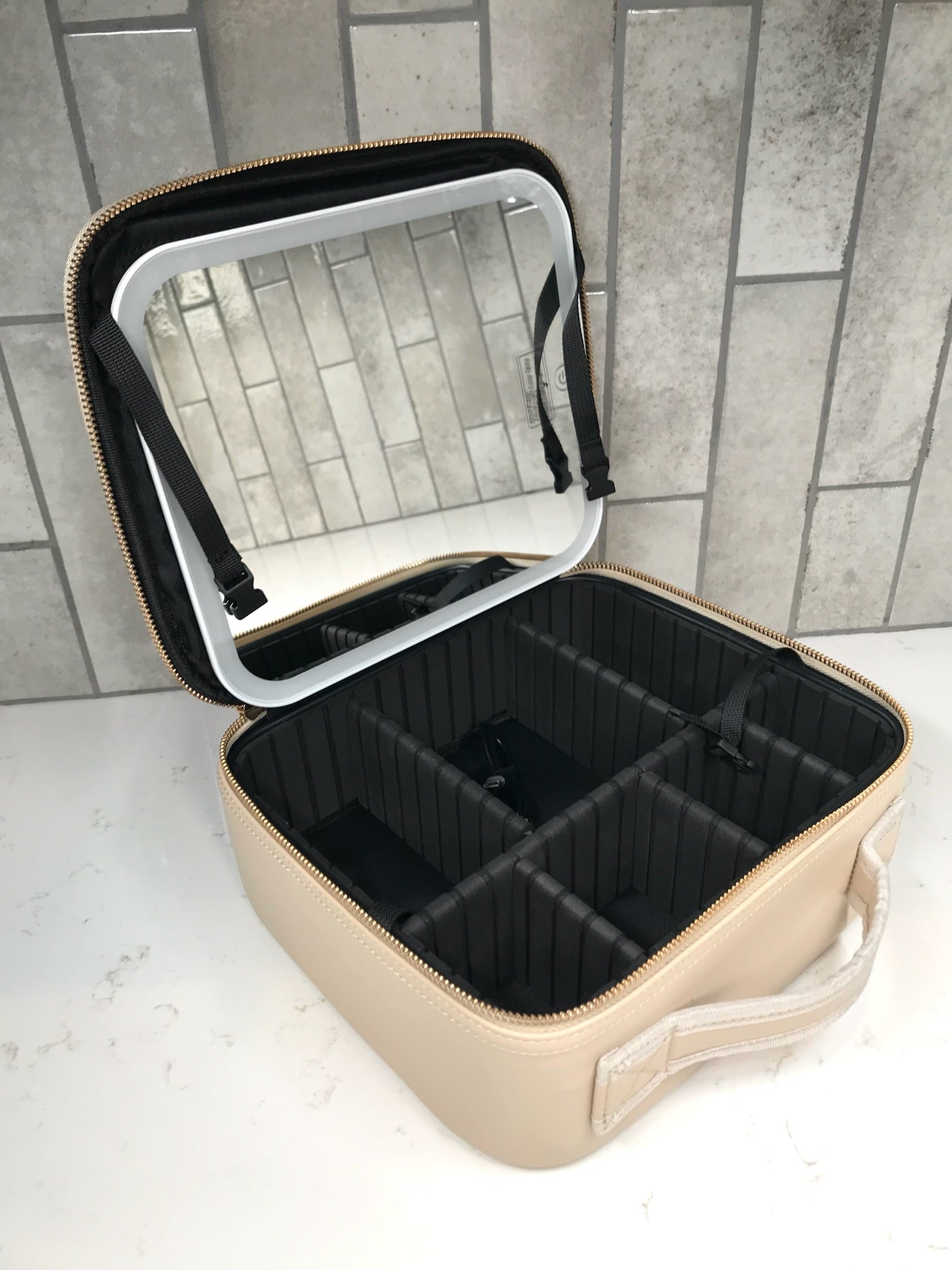 Beige travel cosmetic case with black interior compartments on a tiled floor.