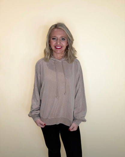 Woman wearing a relaxed fit taupe hooded sweatshirt with drawstrings and black pants, standing and smiling in front of a cream background.