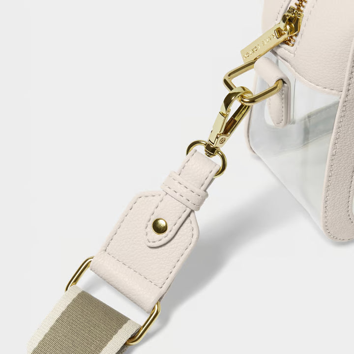 Close-up detail of a handbag strap attachment with a light beige leather strap, gold-tone hardware, and a clear plastic panel on a white background.