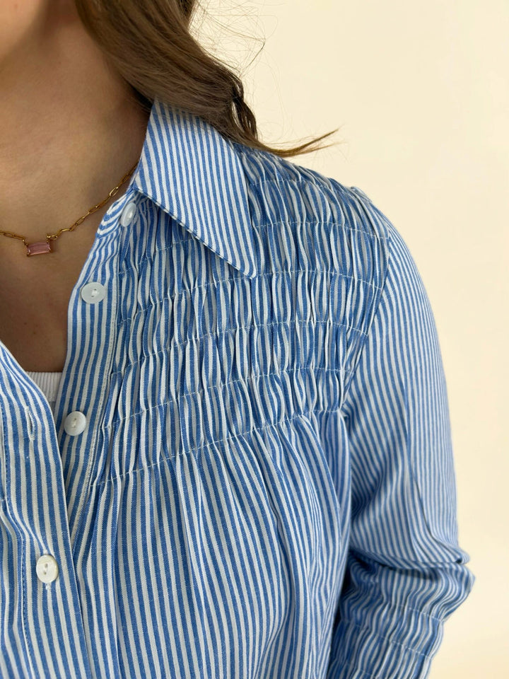Close up of blue striped shirt dress featuring button front, collar neckline, and smocked chest detail