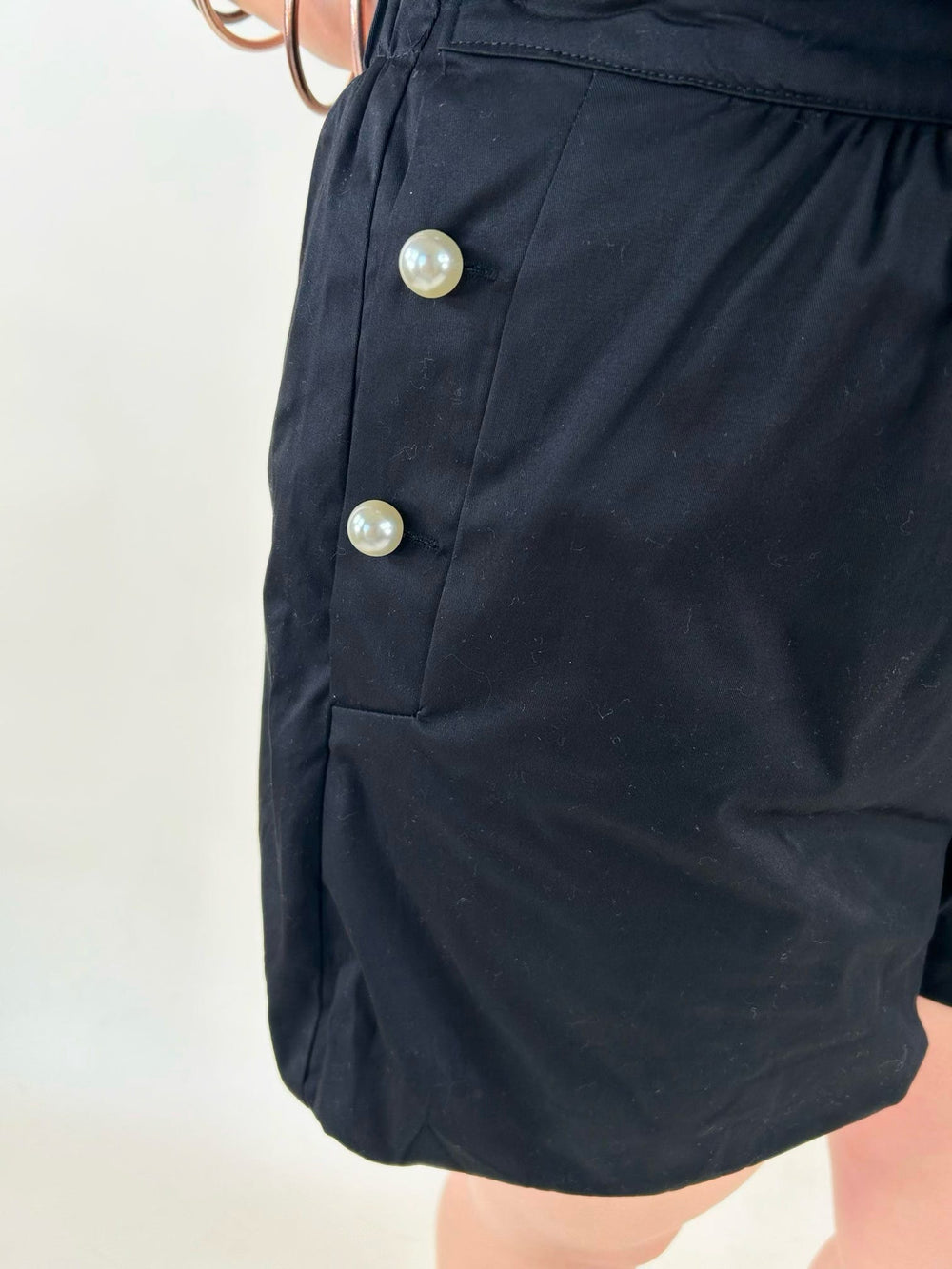 Close-up of black bubble shorts with decorative pearl button side accents, tailored pleats, and modern boutique styling