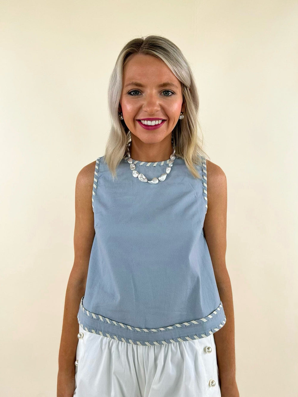Sleeveless light blue top with white whip stitch trim, paired with white high-waisted pants, worn by a smiling model.
