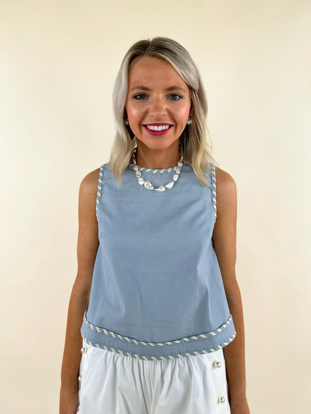 Sleeveless light blue top with white whip stitch trim, paired with white high-waisted pants, worn by a smiling model.
