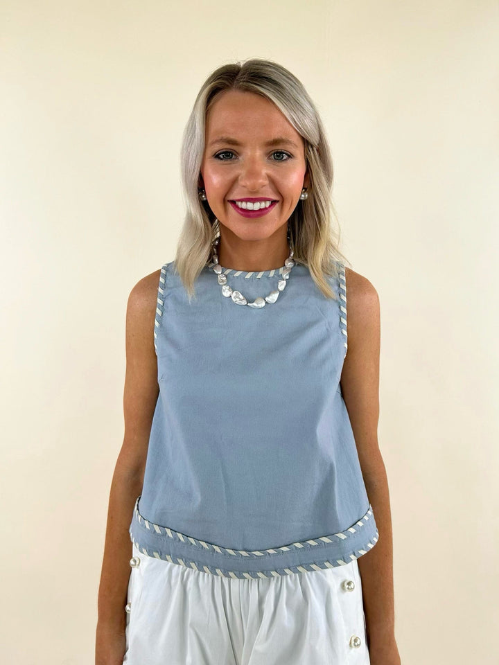 Sleeveless light blue top with white whip stitch trim, paired with white high-waisted pants, worn by a smiling model.