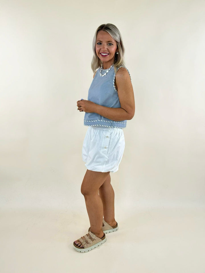 Sleeveless blue tank top with white whip stitch detail, paired with white shorts and beige sandals.